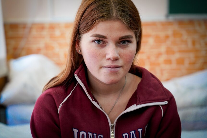 A young redheaded woman in a hoodie sits on a bed