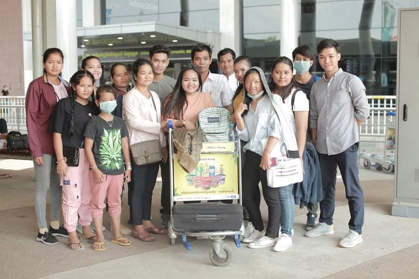 A Cambodian family at the airport, with a girl leaning on a trolley with a suitcase on it.