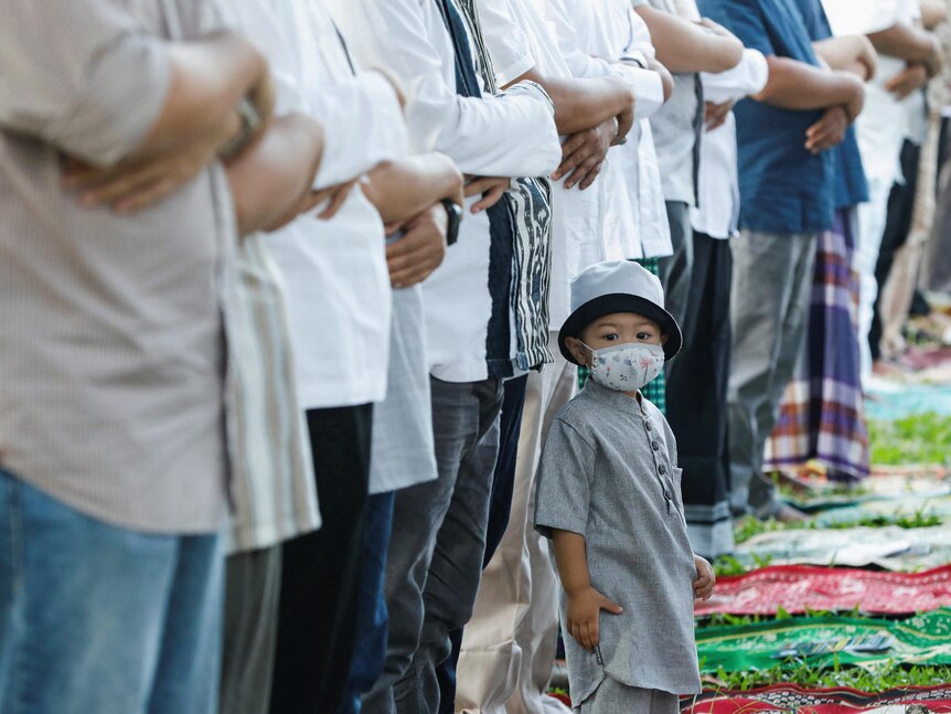 A boy wearing a mask faces the camera as he stands in front of a line of men doing the muslim communal prayer outdoors