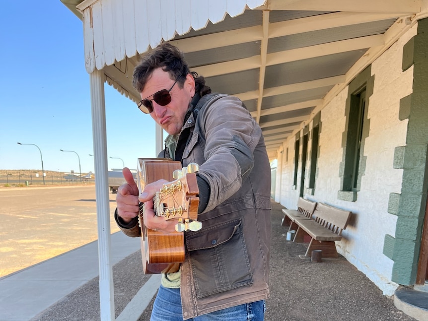 A man in jeans, jacket and sunglasses holds a guitar.