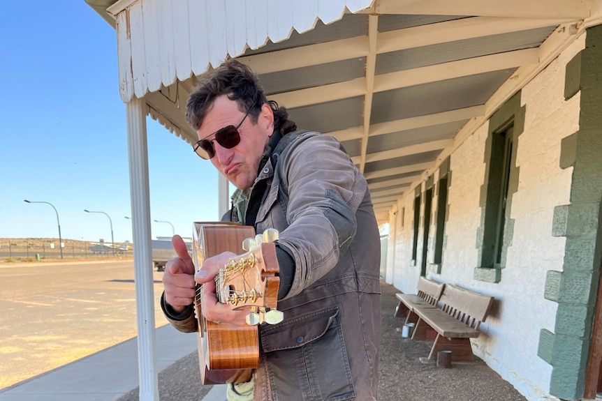 A man in jeans, jacket and sunglasses holds a guitar.