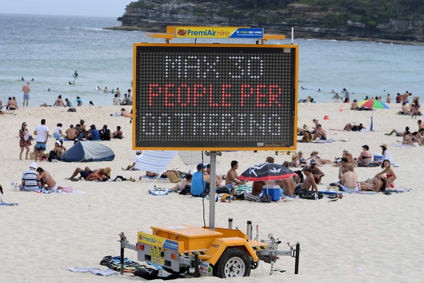 people gathered on a beach behind an electronic sign reading max 30 people per gathering