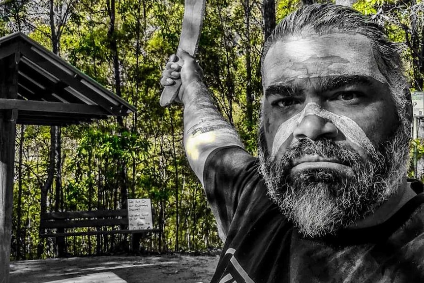 An indigenous man with face paint hold a boomerang in front of bushland