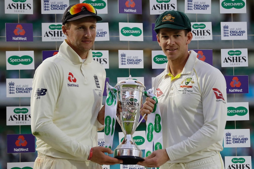 Joe Root and Tim Paine smile awkwardly while holding a large silver trophy.