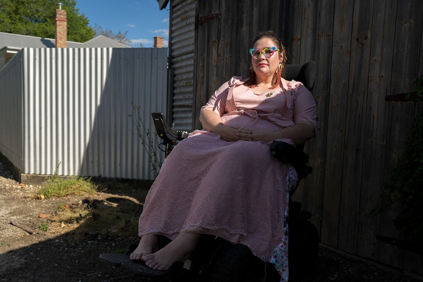 Renay, a disabled writer and artist, wearing a long pink dress, and large rainbow glasses sits in her power wheelchair. 