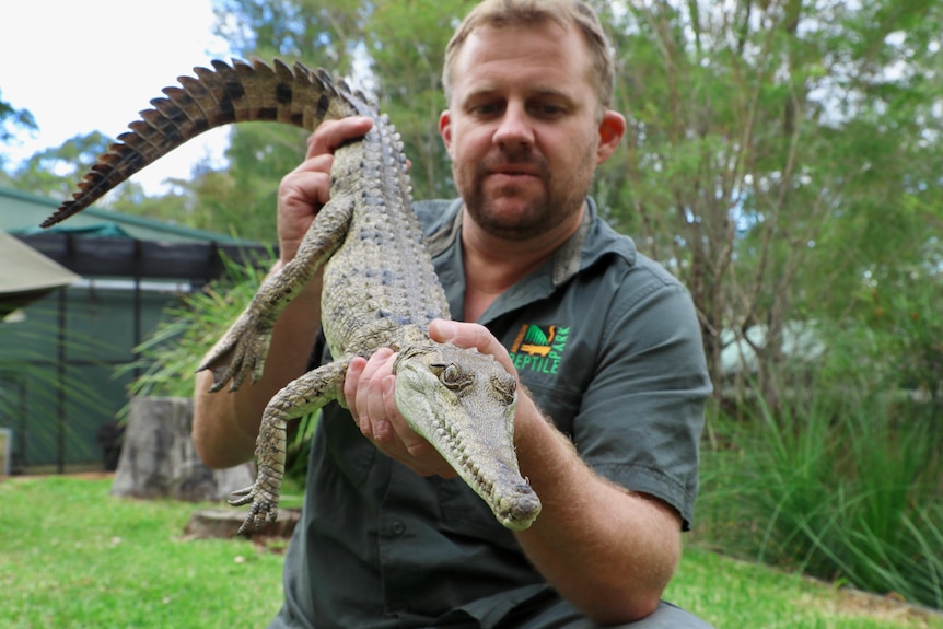 A man with short hair and stubble holds a small crocodile in a backyard.