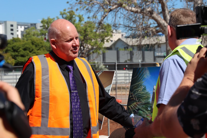 A bald man in an orange high vis vest speaks to several other people at a construction site on a sunny day.