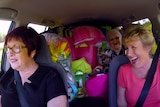 Three women in a car full of luggage and random items, laughing heavily. 
