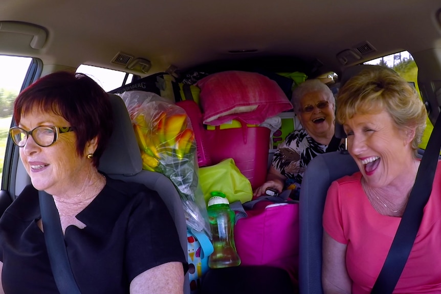 Three women in a car full of luggage and random items, laughing heavily. 
