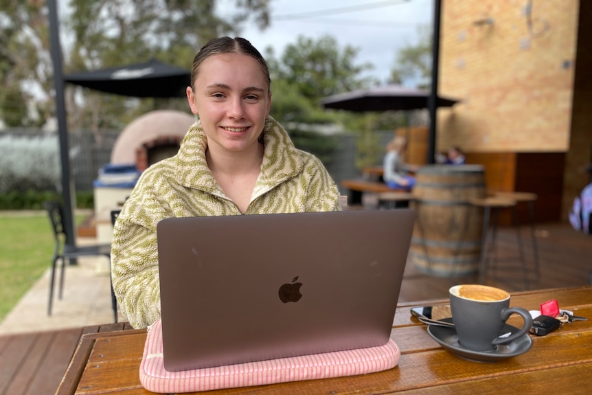 A young woman smiles, blonde slick hair. She sits at wooden table with coffee and laptop. She wears a green/ cream striped jumpe