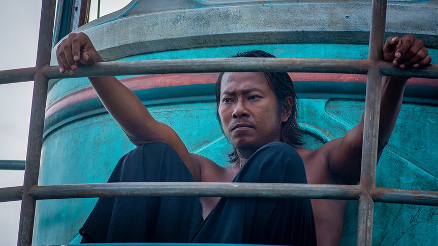 A man with serious expression sits with no shirt on worn-out fishing boat with fading blue paint, hands resting on boat railing.