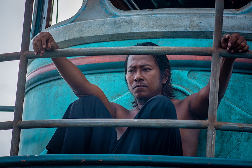A man with serious expression sits with no shirt on worn-out fishing boat with fading blue paint, hands resting on boat railing.