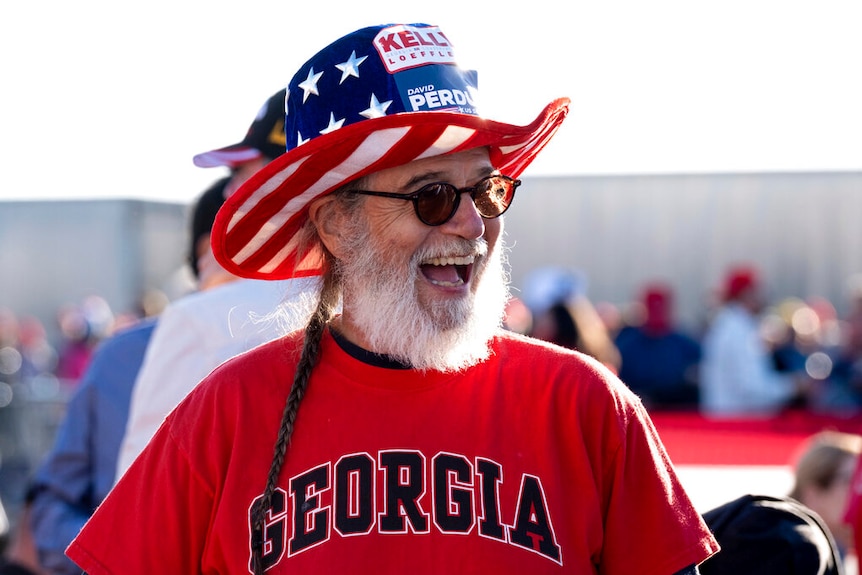 You view a man with a ponytail smiling while wearing sunglasses and a bright red shirt that reads 'Georgia'.