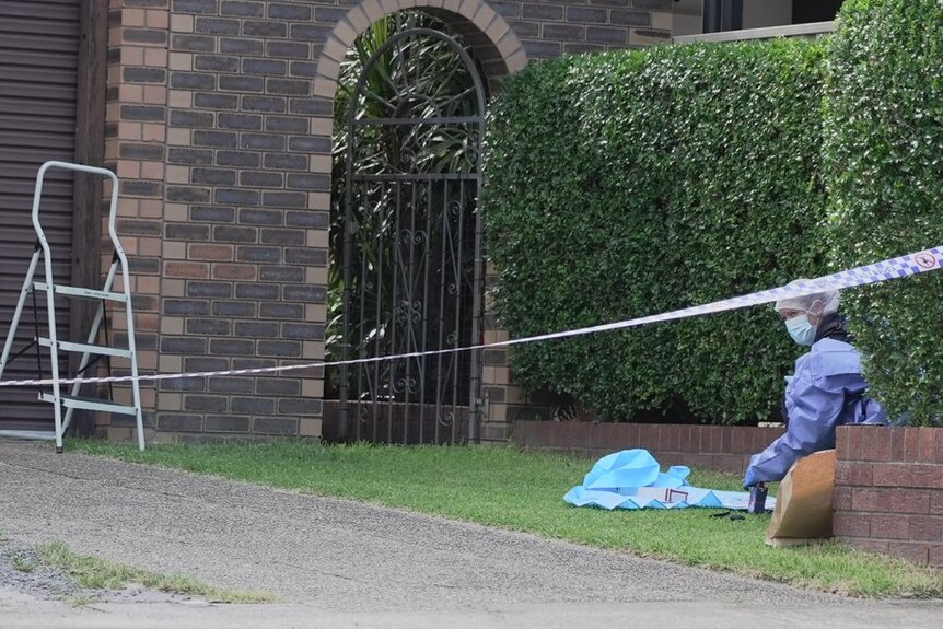 A woman in blue scrubs and a mask examing evidence in the front garden with police tape cordoning off the house