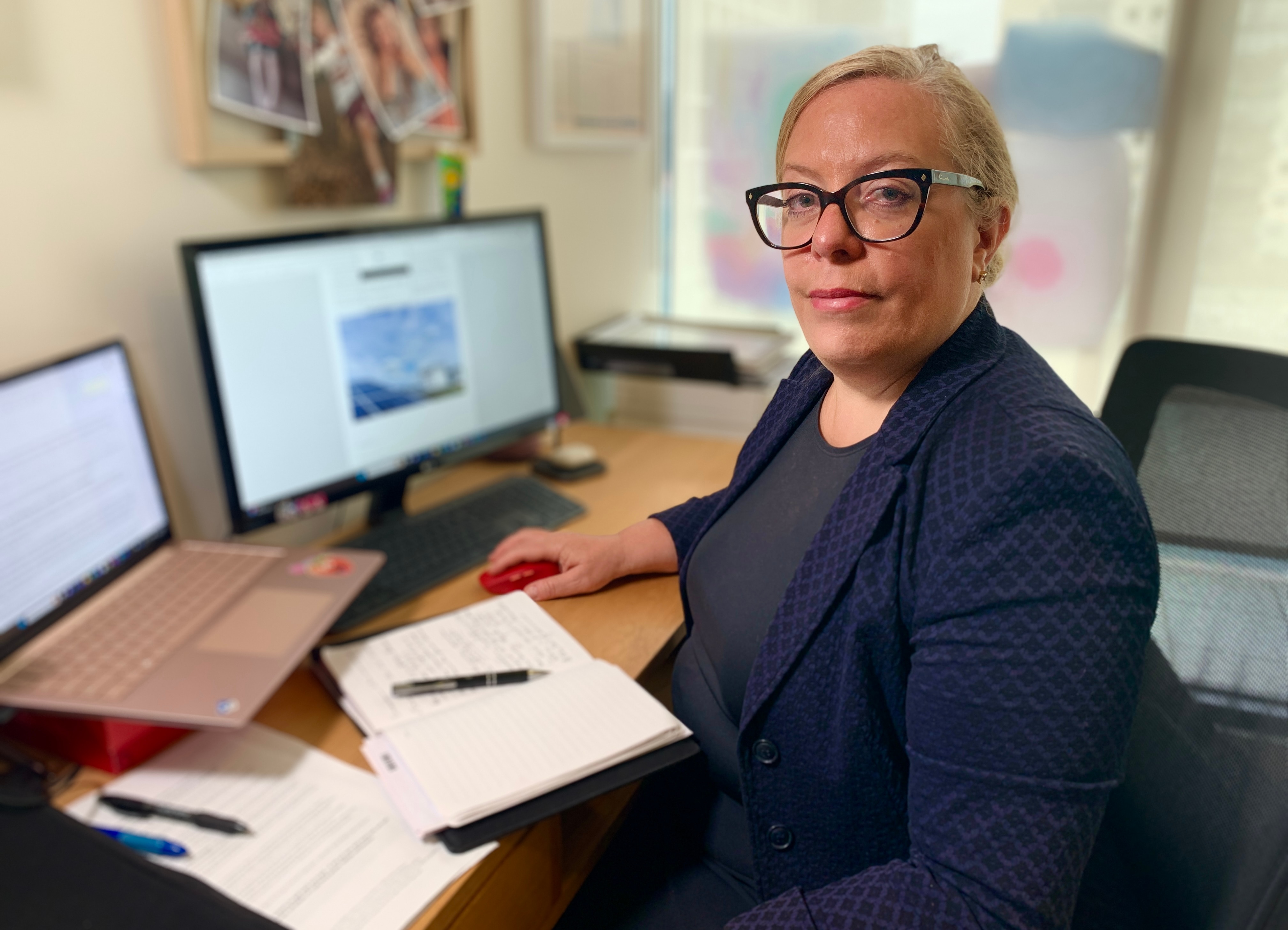 Una mujer está sentada frente a su computadora, con su trabajo frente a ella. Está mirando a la cámara.
