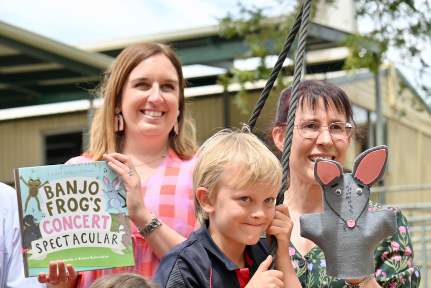 A woman holds a picture book next to another with a hand puppet of a bilby with a school child on play equipment between them.