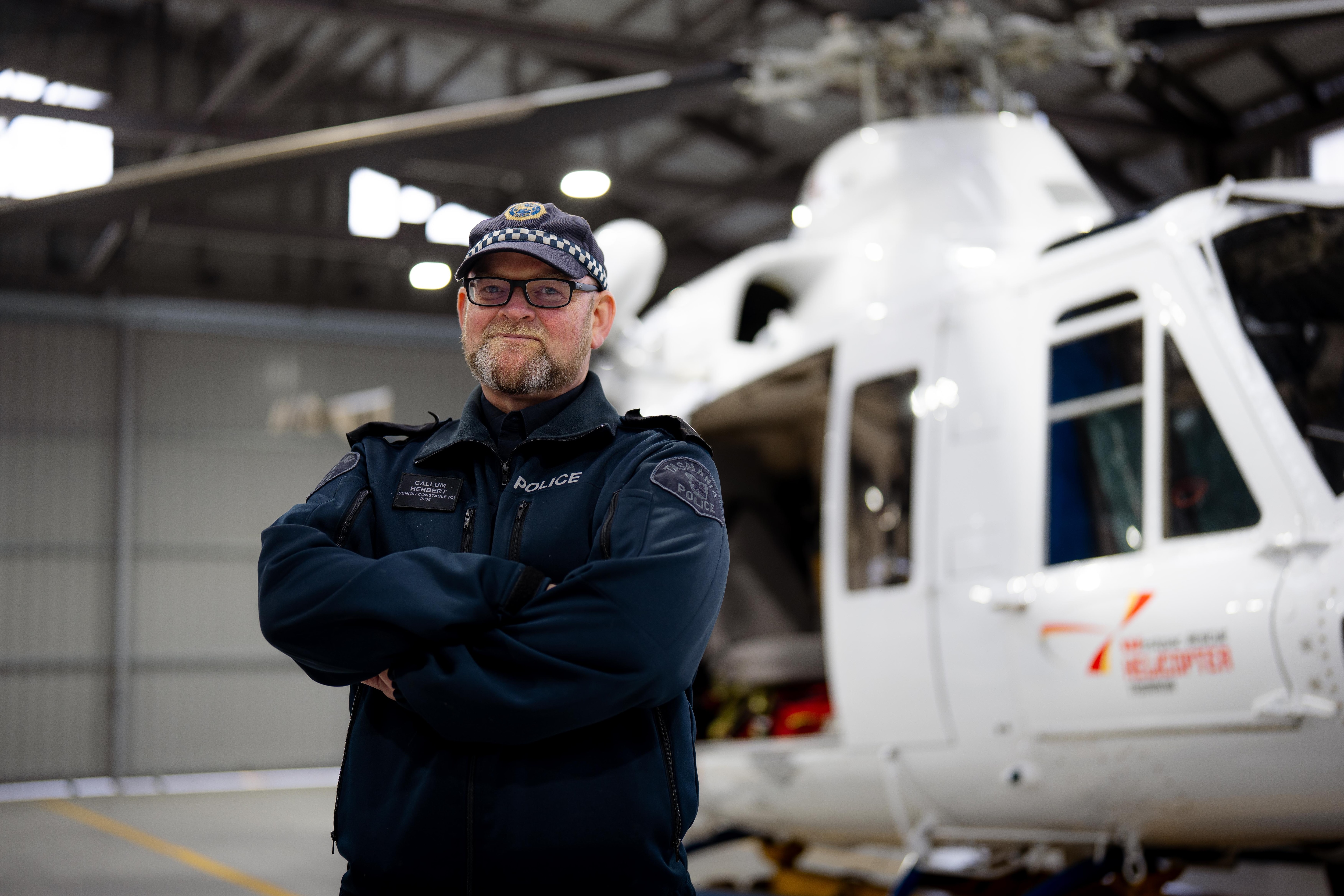 Callum Herbert, wearing police uniform, crosses his arms posing for a photo with a white rescue helicopter behind him.