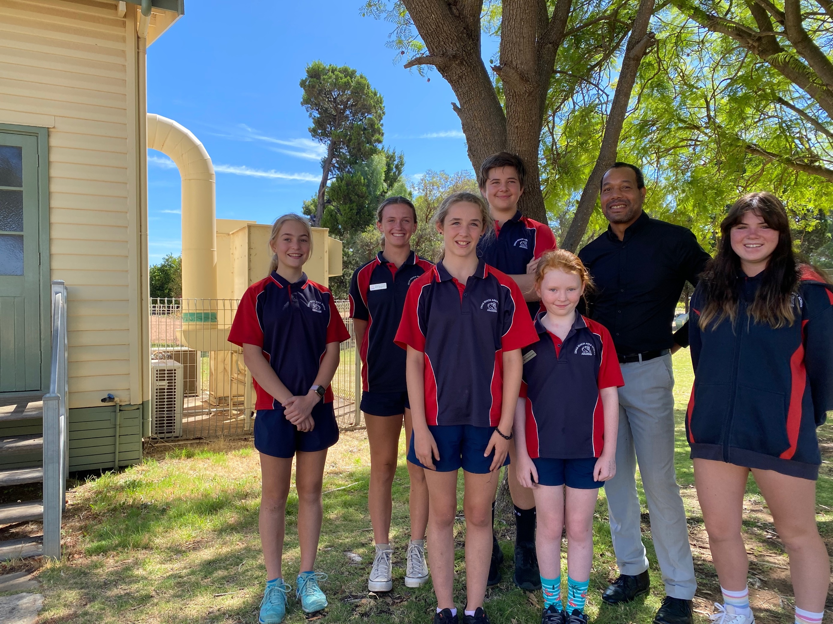 six school children wear red and blue navy uniforms, stand with their principal in the shade of a tree