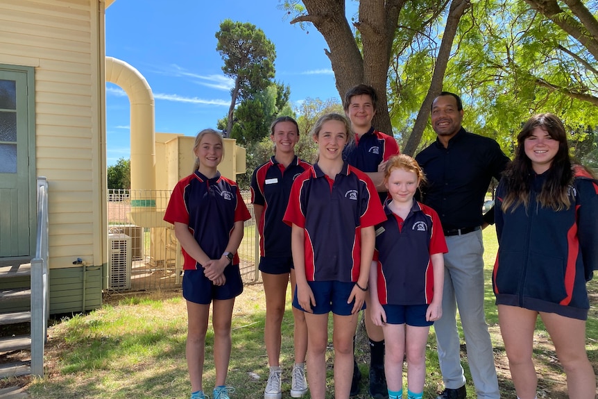 six school children wear red and blue navy uniforms, stand with their principal in the shade of a tree