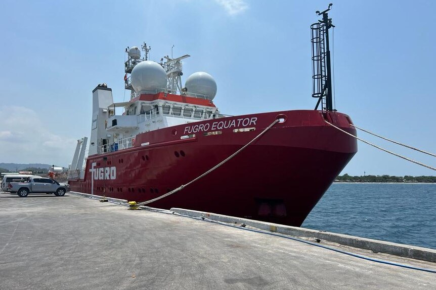 A large survey vessel with a red hull sits at dock.