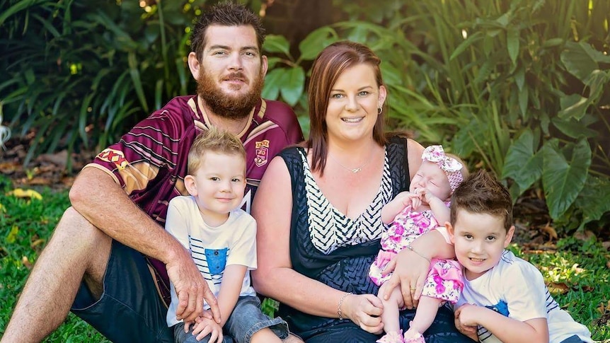 Family of mum, dad, and three small children sitting on the grass smiling.