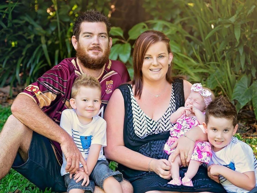 Family of mum, dad, and three small children sitting on the grass smiling.