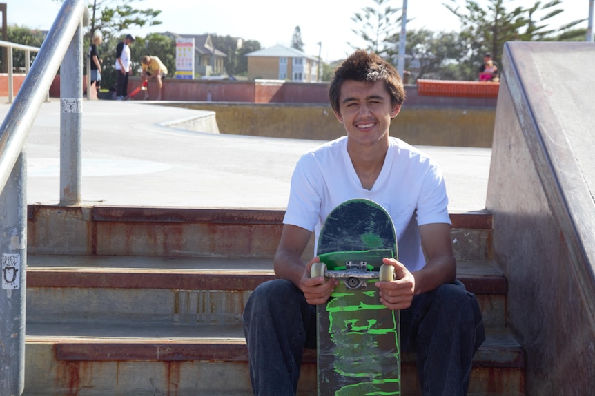A young man in a white shirt sits on concrete steps in front of a skate bowl smiling. He is holding his skateboard in his hands