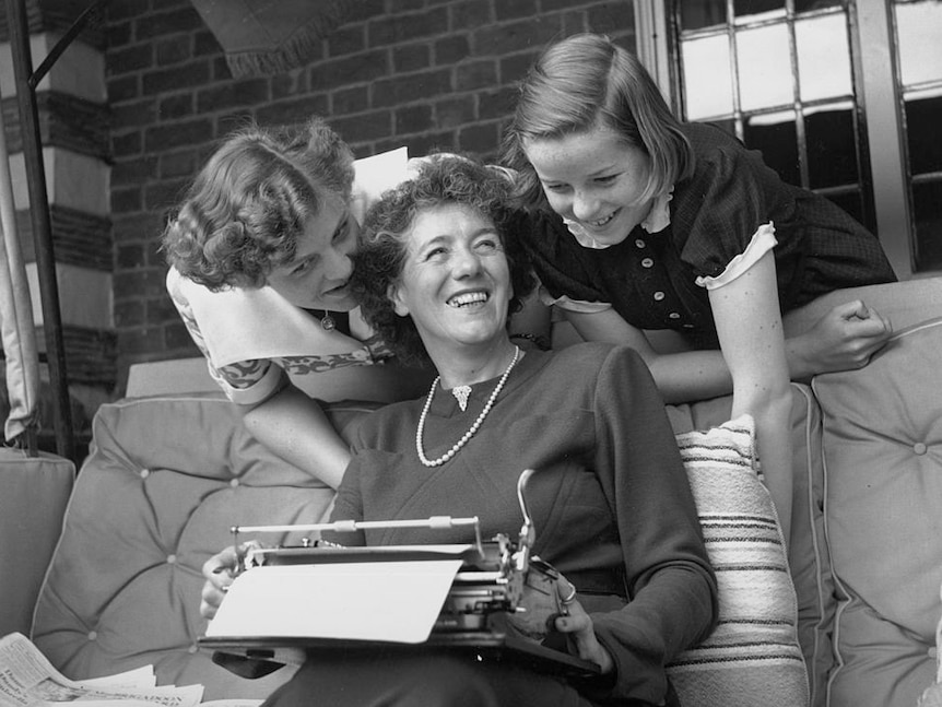 1949 photo of Enid Blyton smiling, holding typewriter in lap. Her two daughters Gillian and Imogen lean over her, smiling.