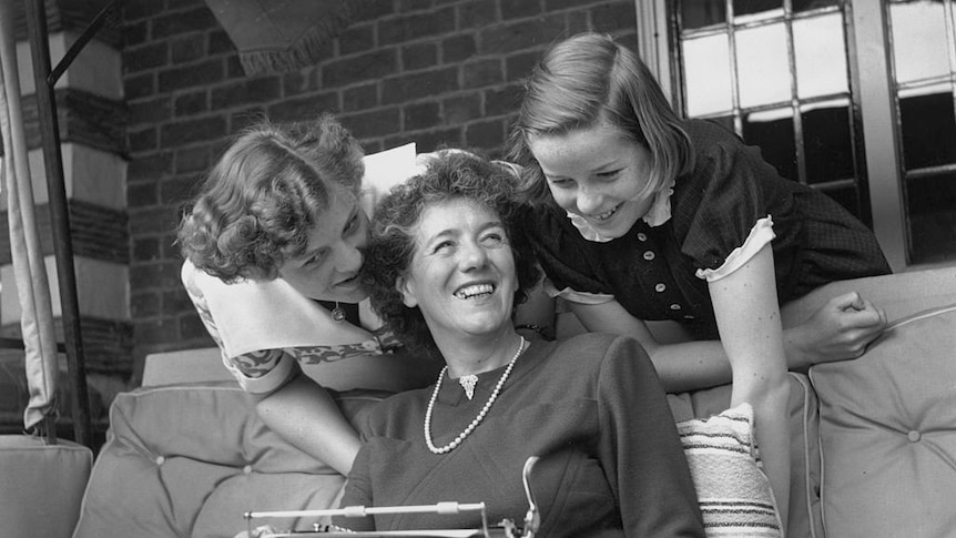 1949 photo of Enid Blyton smiling, holding typewriter in lap. Her two daughters Gillian and Imogen lean over her, smiling.