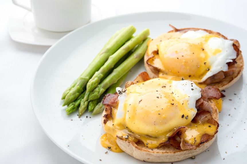 Hollandaise sauce poured over poached eggs, bacon and English muffin with asparagus on the side.