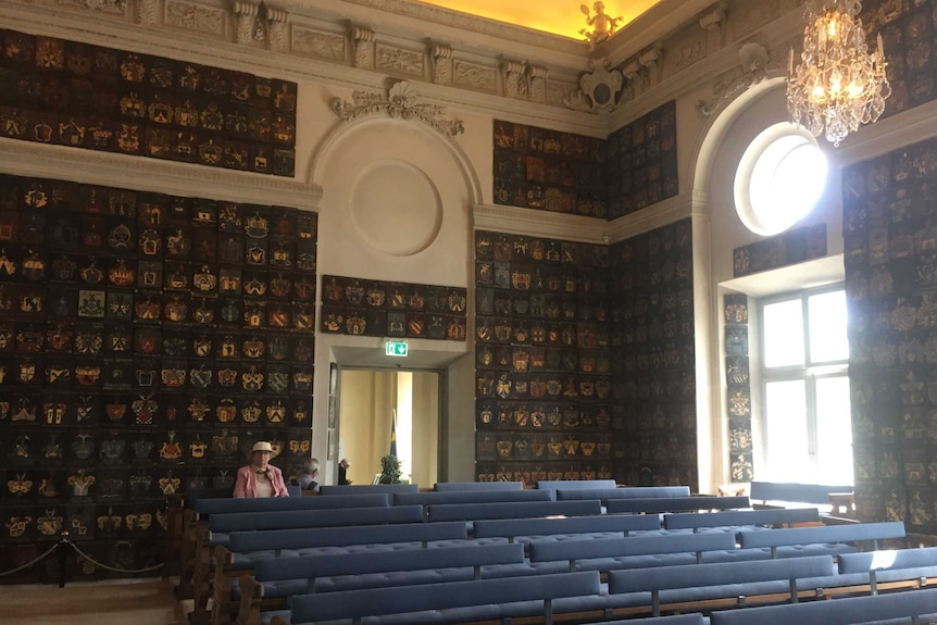 Hundreds of coats of arms line the walls of an ornate room. A woman sits in the last row of a number of benches in the room.