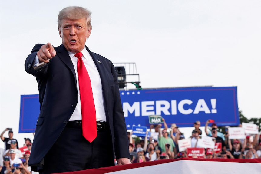 Donald Trump stands in front of a campaign sign, pointing at the camera.