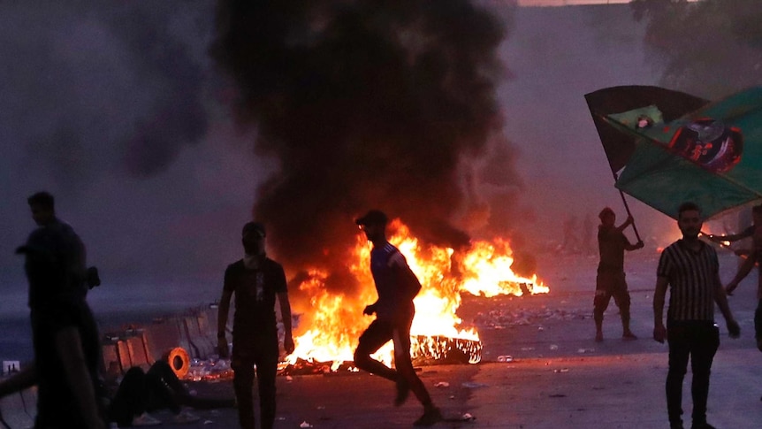 Anti-government protesters set fires as they rally on a closed Baghdad street. Smoke fills the air.