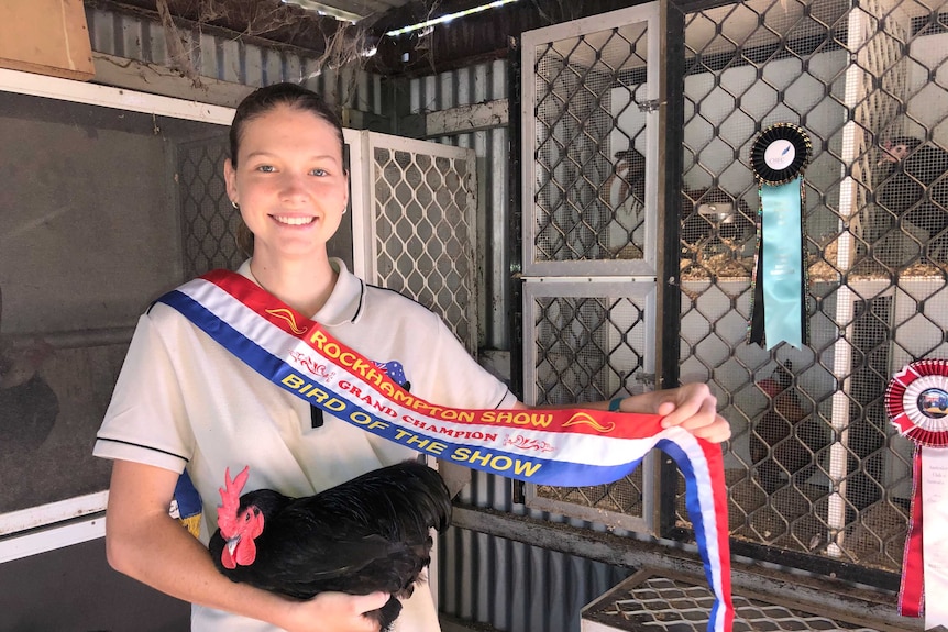 A young woman stands holding a black chicken. She is smiling and has a winning ribbon saying 'Grand champion bird of the show'.