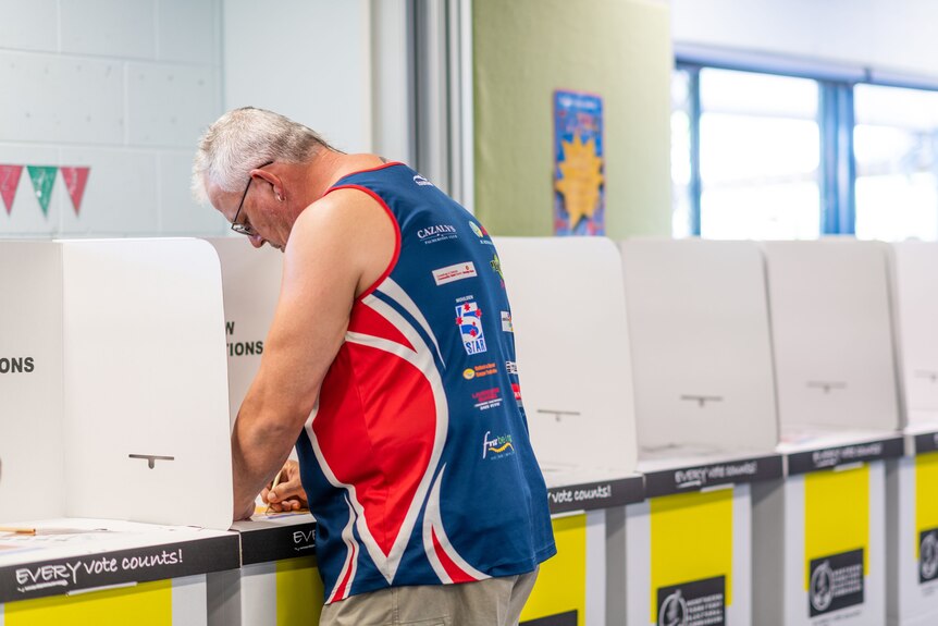A man standing in a cardboard polling booth, writing on his ballot paper.