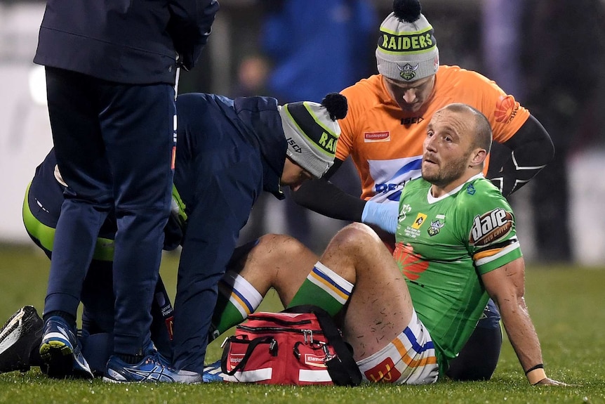 Josh Hodgson sits on the ground with people around him holding and looking at his knee