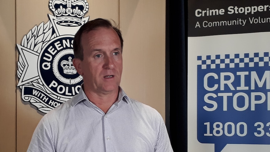 a man in a white shirt addresses media in front of a Queensland Police symbol