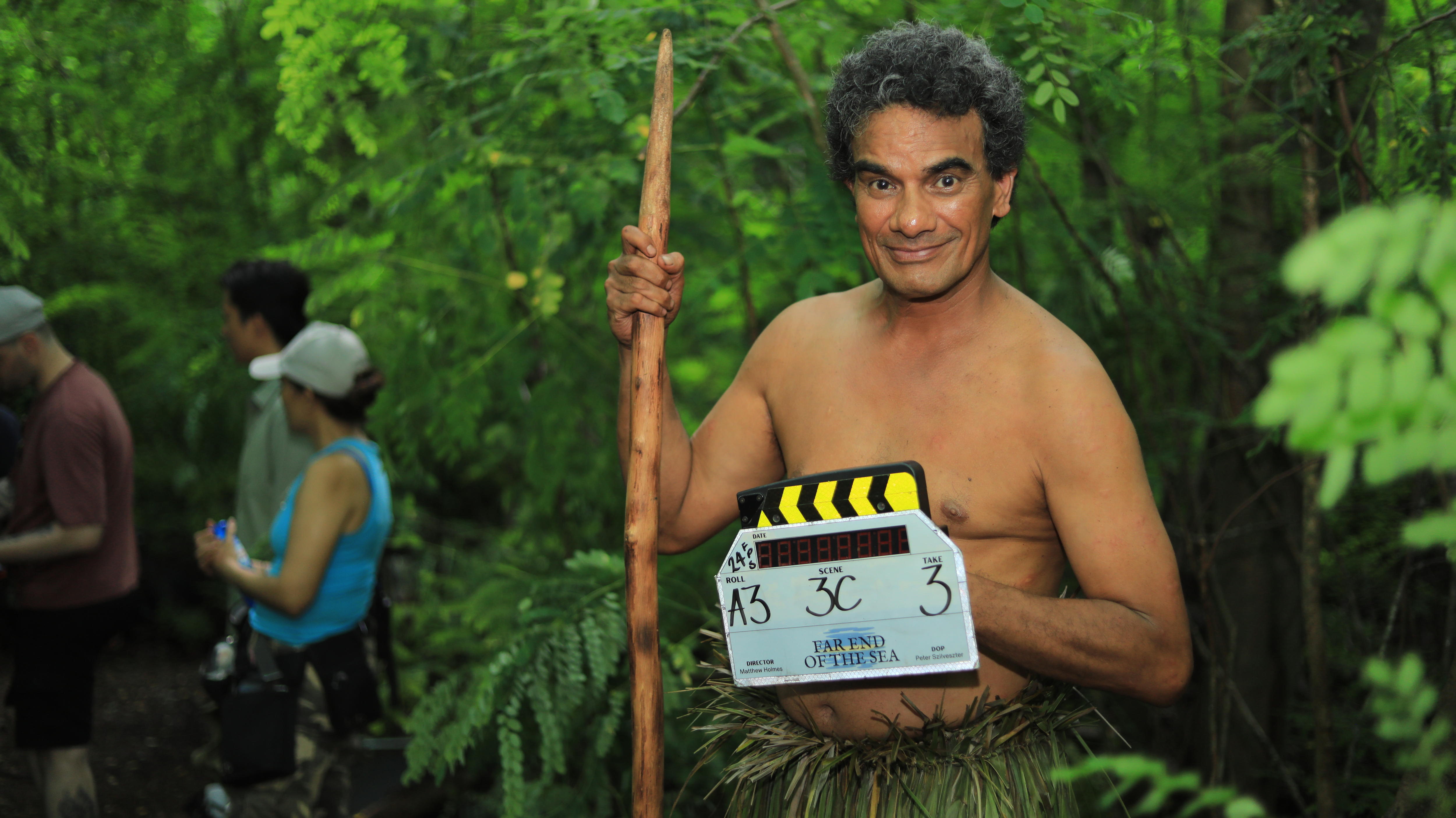 A man wearing traditional wear smiles and holds a clapperboard in the jungle.
