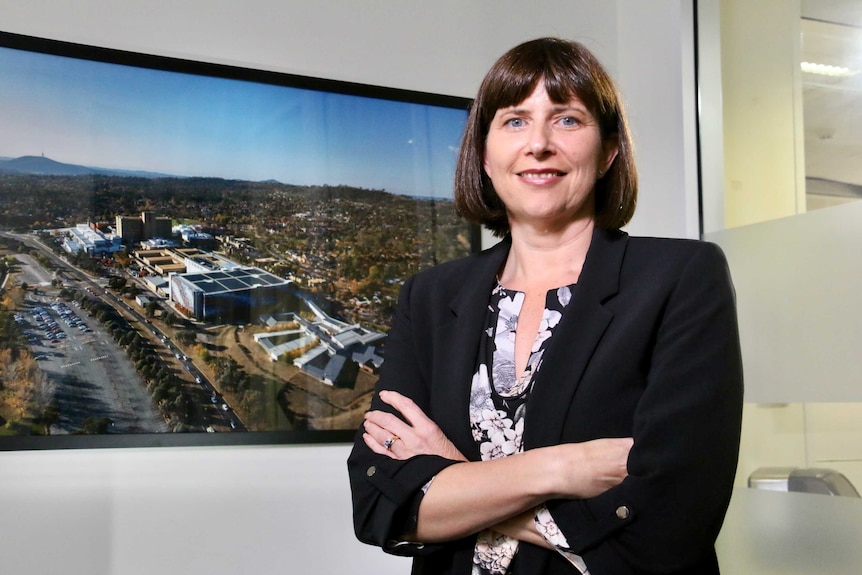 A woman stands with crossed arms but smiling in front of an aerial photo of the Canberra Hospital.
