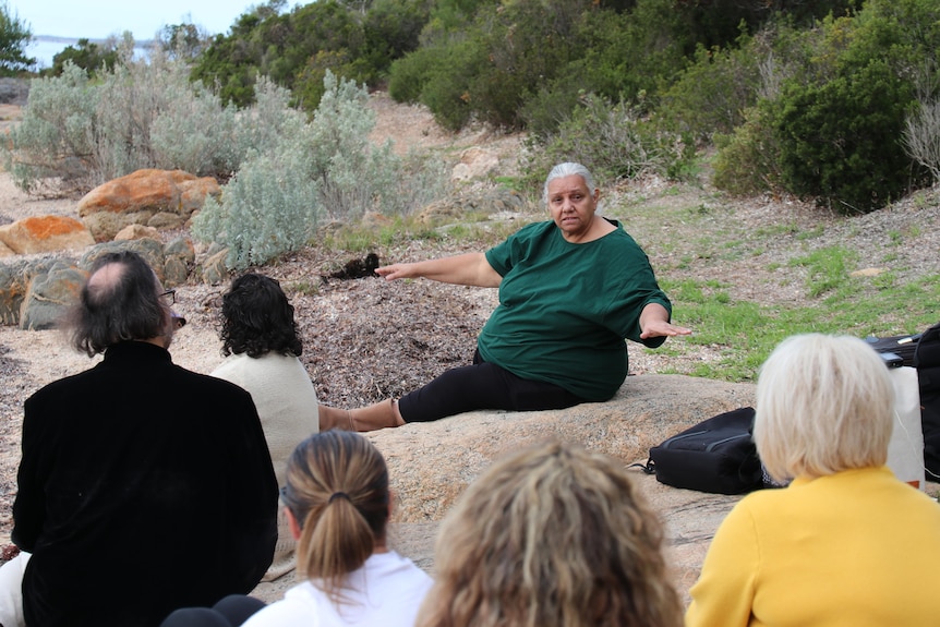 Indigenous woman on a rock with arms out by her sides talking to small gathering of people, backs of heads in photo