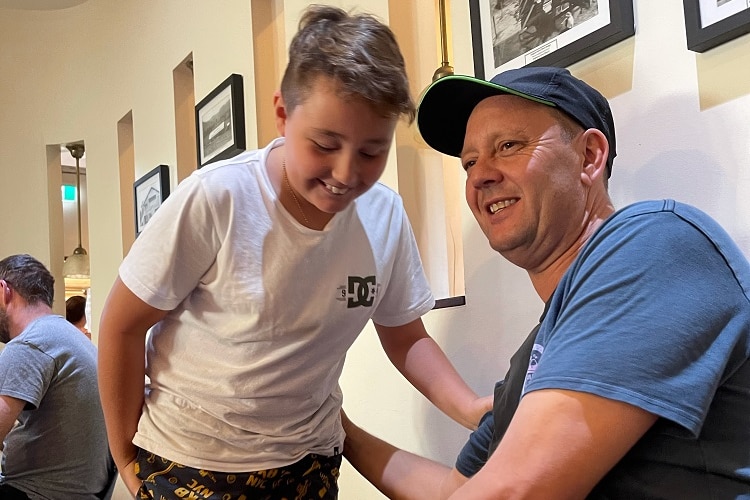 A dad wearing cap smiles as his young son leans in to talk to him, smiling.