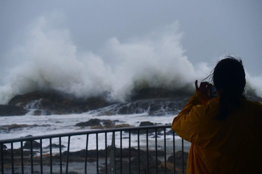 The back of a woman taking a photo of large wave crashing against rocks.