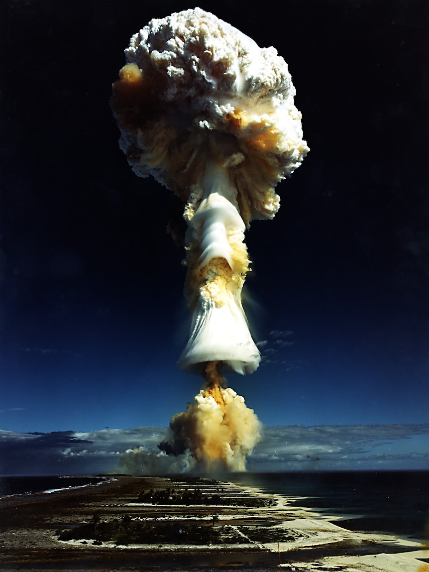 A film photo shows a milky-yellow mushroom cloud shoot up from low-lying coral atolls.