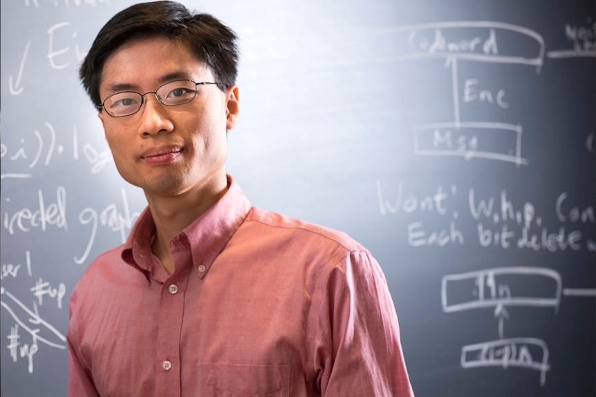 A man in a pink button-up shirt stands in front of a blackboard with equations written out