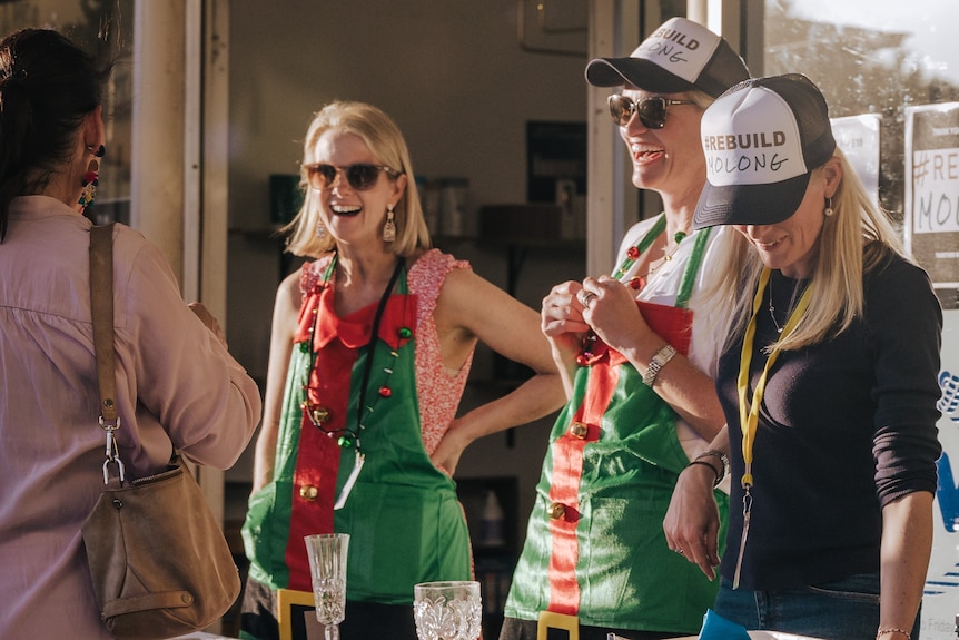 Three women with blonde hair laugh as they serve a customer while wearing Christmas aprons 