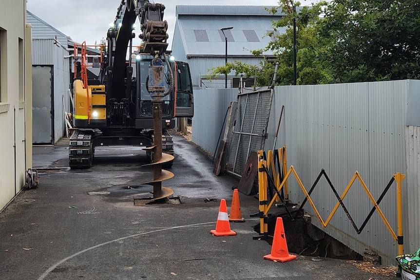 A large machine drills down into a driveway, with a portion of a sinkhole extending under a fence nearby.
