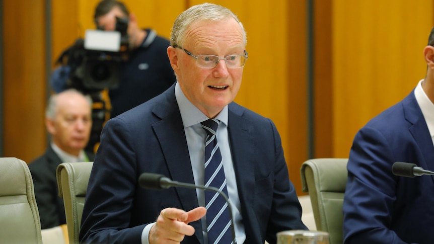 Philip Lowe wearing glasses and a suit sits at a table and speaks.