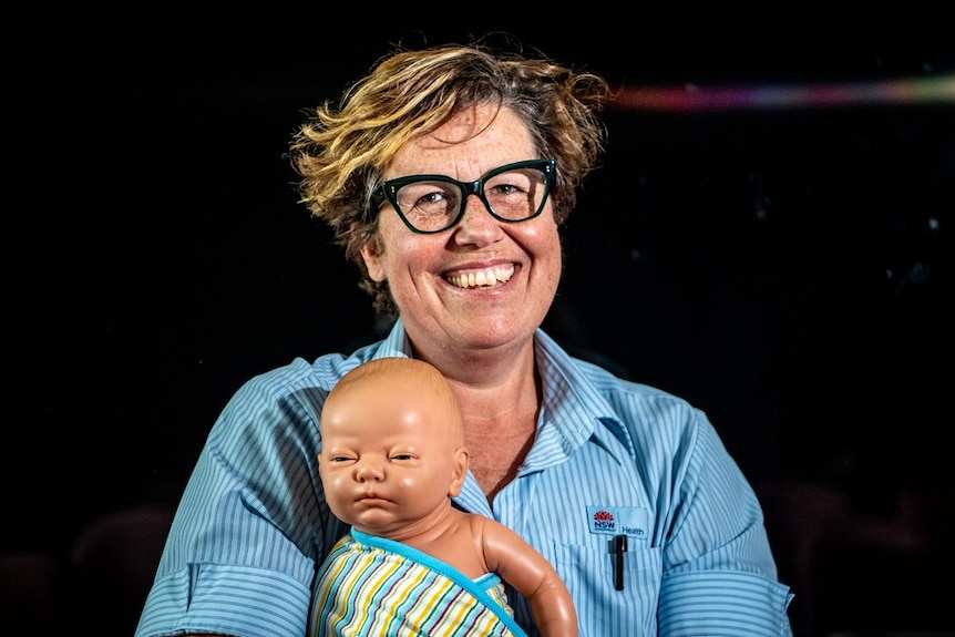 Midwife Cherie holds a plastic doll and wears her blue work uniform. She's smiling. 