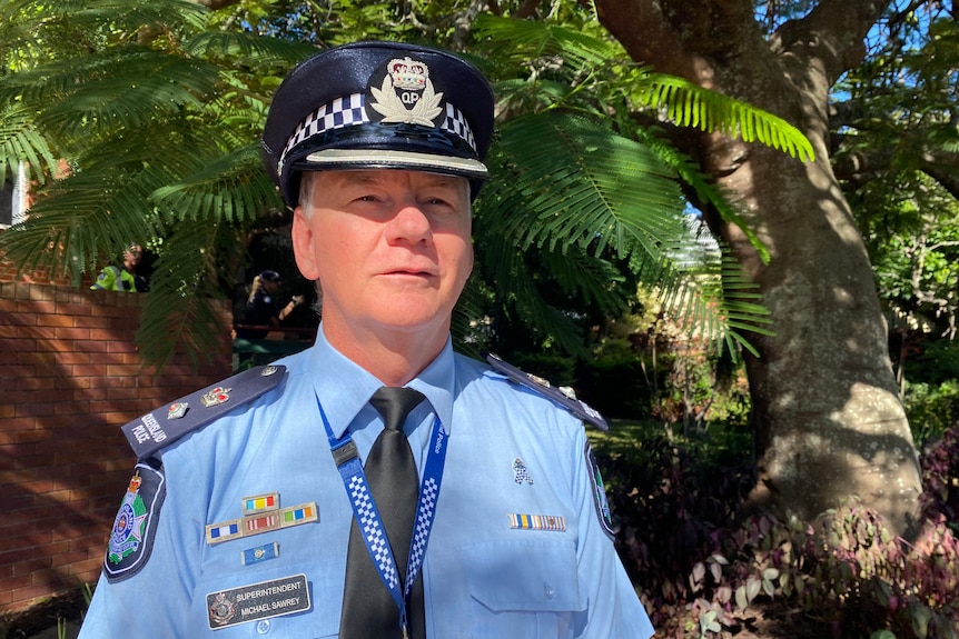 A senior policeman stands in uniform looking serious with a background of trees.