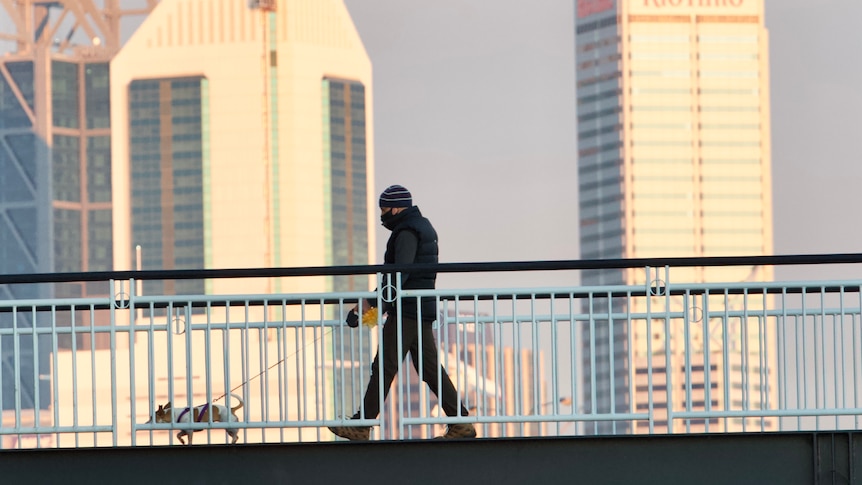 A masked man walks his dog along the footbridge at Claisebrook Cove with the Perth skyline in the background